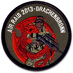 Ecusson rond brodé de la base aérienne 901 de Drachenbronn. L'écusson est brodé sur une toile vert kaki, il représente un militaire armé entremêlé avec un dragon rouge. Au dessus du dessin, il est écrit Air raid 2013 - Drachebronn.