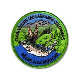 Ecusson rond brodé du club de pêche à la mouche de Colomiers. Il représente une mouche de pêche rose et verte, devant une eau bleue et un paysage de montagne verdoyant. Sur le heut de l'écusson, le texte club loisirs Leo lagrange colomiers est brodé, sur le bas le texte Pêche à la mouche. La bordure de l'écusson est verte.