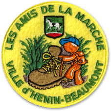 Ecusson rond brodé du club de randonnée des amis de la marche à Henin beaumont. L'écusson a un fond jaune sur lequel une chaussure de montagne marron est représentée avec un petit personnage de randonneur orange à côté. derrière, une touffe d'herbe verte est brodée. Au-dessus du dessin, il y a le blason de la ville d'Henin beaumont. En haut de l'écusson, le texte les amis de la marche et en bas, le texte Ville d'Henin beaumont.