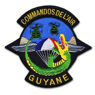 Acusson brodé de l'armée de l'air. Il représente un parachute aux couleurs du drapeau tricolore, derrière un varan vert et jaune, et sous deux hélicoptères verts kaki. Deux bandeaux noirs, en haut et en bas de l'écusson en forme de cercle, où le texte suivant est brodé : commandos de l'air, guyane.