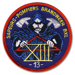 Ecusson rond, brodé des sapeurs pompiers de Bruxelles. il représente, sur un fond bleu foncé, un homme obscur portant un triangle rouge dans ses mains et le chiffre XIII en lettres d'or. Dans son dos, deux haches dépassent. Au dessus, le texte Sapeurs pompiers Brandweer BXL est écrit en jaune et en bas, le texte 13 est brodé en blanc.