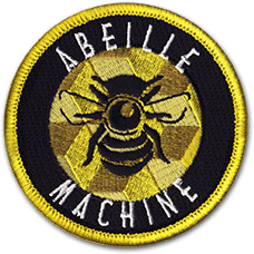 Ecusson brodé de l'association Abeille machine. Il est rond et représente une abeille sur une alvéole de ruche. Le tour du dessin est noir et bordé de jaune. On y lit le texte en blanc abeille machine.