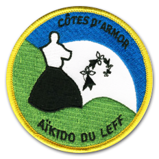Ecusson brodé du club d'aïkido du leff. Il représente un homme en tenue traditionnelle blanche et noire de dos, devant un lever de soleil sur un sol vert. En haut de l'écusson, le texte Cotes d'armor et en bas, le texte Aïkido du leff sont brodés en noir. le bord de l'écusson est jaune.