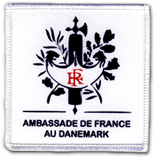 Ecusson imprimé de forme carrée. Sur un fond bleu, le texte ambassade de France au Danemark est écrit en bleu foncé sous le symbole de la république française, dans lequel les lettres RF sont écrites en rouge.