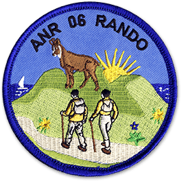 Ecusson rond brodé du club de randonnée ANR 06 rando. Il représente deux randonneurs marchant sur un chemin de montagne. Au-dessus d'eux, un chamois est brodé, et derrière la montagne, un soleil se lève.