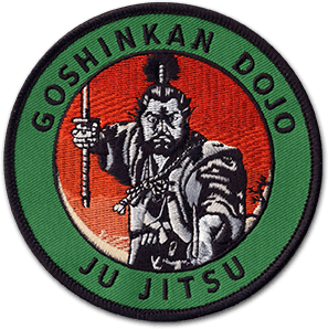 Ecusson rond brodé de l'école de Ju jitsu. Au centre, un samouraï en gris et noir sur fond rouge dégradé. Le contour de l'écusson est vert, dessus il est écrit Goshinkan Dojo, Ju jitsu.
