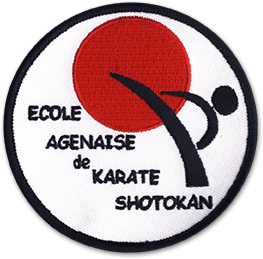 Ecusson de l'école de karaté Shotokan d'Agen. Il est rond, sur fond blanc bordé de noir. Il représente un rond rouge en haut, avec une silhouette d'homme tapant du pied dedans. Le niom de l'école est écrit en noir sur la partie gauche.