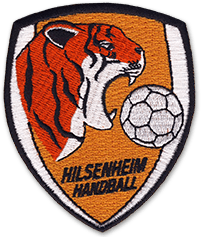 Ecusson du club de handball Hilsenheim. Il est en forme de blason pointe vers le bas, et dessus sont brodés un tigre et un ballon de hand. Sous le dessin, le nom du club est écrit sur fond orange.