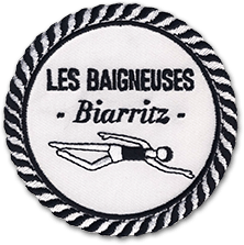 L'écusson brodé rond en noir et blanc représente le logo du restaurant les baigneuses à Biarritz. Sur un fond blanc, une baigneuse est représentée, nageant, de profil. Elle porte un bonnet de bain noir. Au dessus, le nom de l'hôtel restaurant est brodé en noir : les baigneuses, biarritz. Le bord de l'écusson est un bourdon imitant une corde marine noire et blanche.