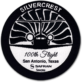Ecusson de Safran pour le 100ème vol du moteur Silvercrest. L'écusson rond brodé et imprimé est en deux couleurs et représente un avion de face, sur un fond de réacteur. Sous l'avion, le texte 100th flight, San Antonio, Texas, Safran.