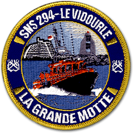 Ecusson brodé rond des sauveteurs en mer de la grande Motte. Il représente un bateau rouge naviguant sur la mer, devant un port. En haut de l'écusson on lit SNS 294 - le Vidourle et en bas, la grande motte.