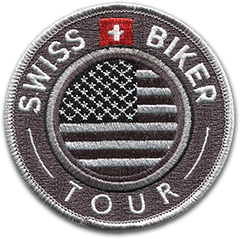 Ecusson du club de motards swiss biker tour qui organise des circuits à moto aux Etats unis. Il est rond et reorésente, en gris clair sur gris foncé, une partie du drapeau des Etats Unis. Autour, un cercle gris clair puis le texte Swiss Biker Tour en gris clair, avec le drapeau suisse en rouge et blanc.
