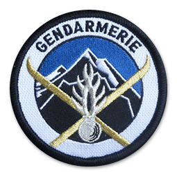 Ecusson brodé rond des unités de montagne de la Gendarmerie Nationale. Il représente une montagne noire et blanche sur un fond de ciel bleu. Devant, deux skis croisés couleur or et une grenade à Huit branches. Autour, il y a un bandeau blanc sur lequel il est écrit Gendarmerie en lettres capitales.